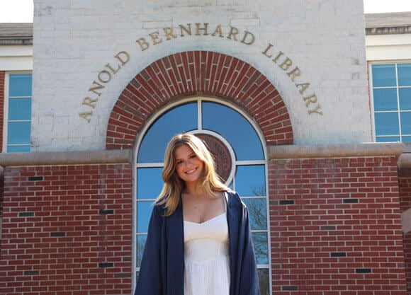 Hannah Jack smiles in a white dress and gown in front of the library.
