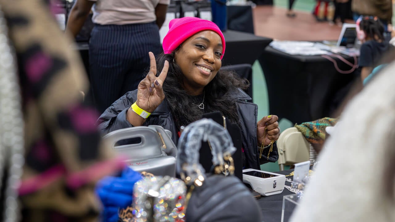 A jewelry vendor wearing a hot pink beanie gives a peace sign with their fingers.