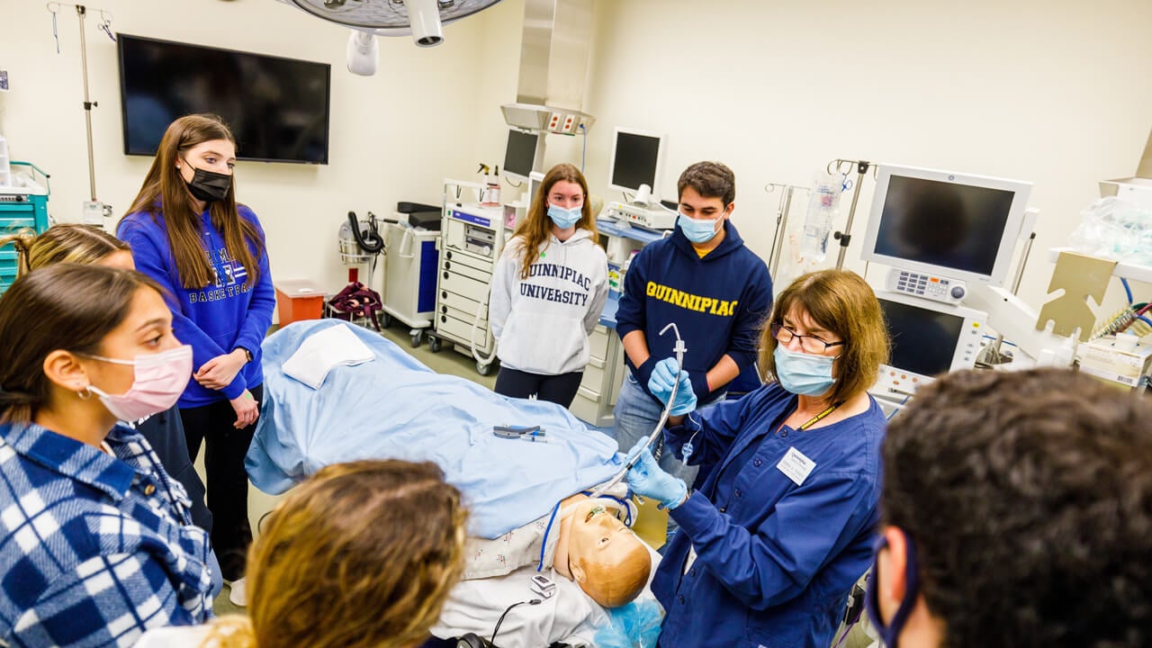 Half a dozen students and a professor practice clinical skills with a mannikin
