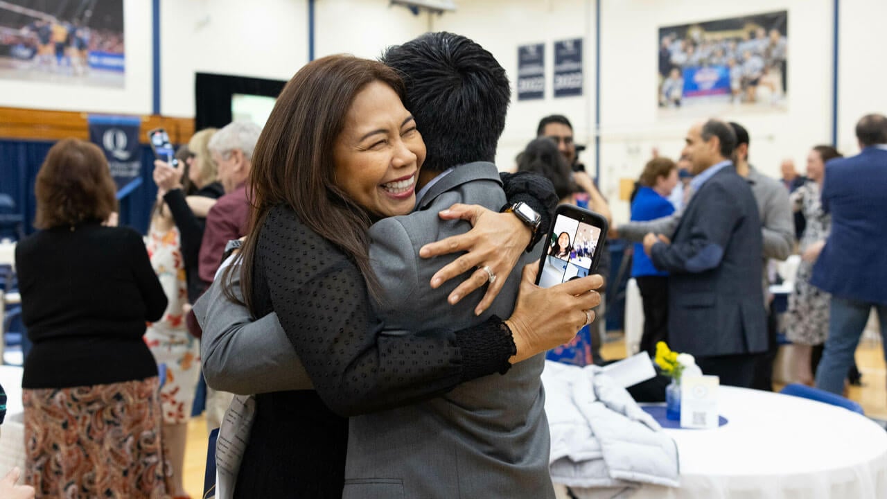 Parent hugs their student with a wide smile on their face holding a phone on a video call with family