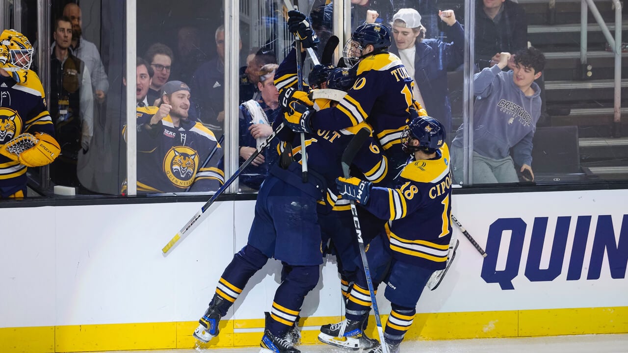 Quinnipiac men's ice hockey players group hug in celebration as fans cheer in the stands