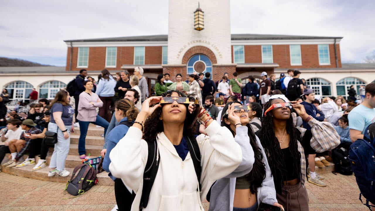 Students hold up solar eclipse glasses to their eyes in front of the library.