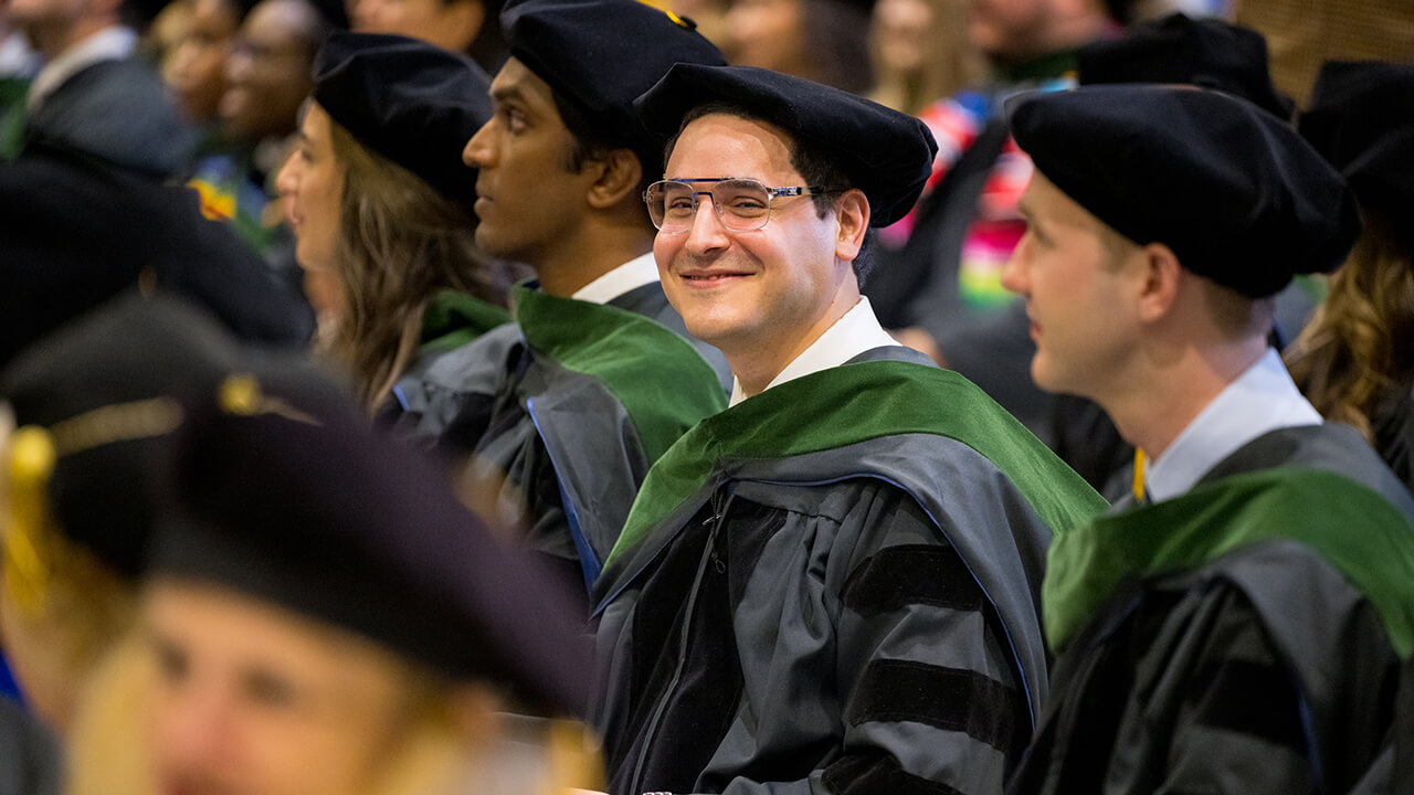 Graduate in glasses sitting in row smiles for camera