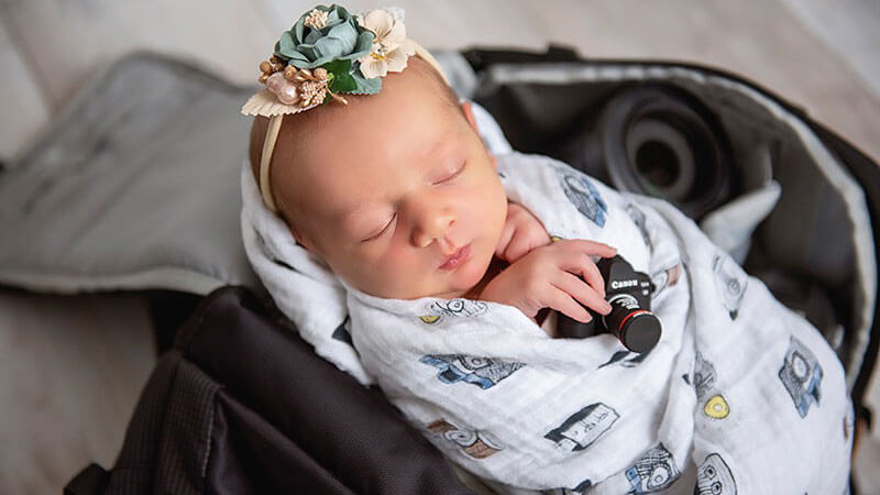 Infant Nora Schussler swaddled in a blanket holds a toy camera