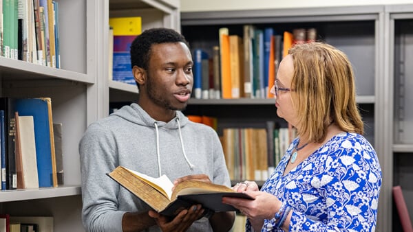 An international student holds a book and speaks with a staff member in a library