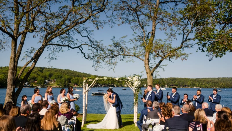 April Simell and Sean Sweeney kiss under a trellis in front of a lake while guests clap