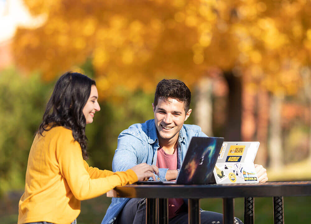 Two students sit at an outdoor table with their laptops with yellow fall foliage in the background