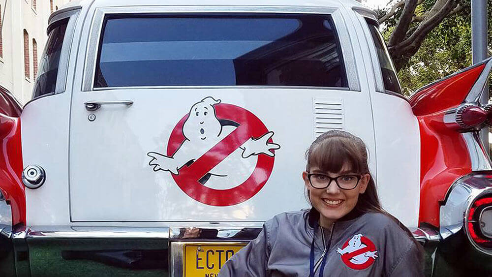 Brooke Mommsen poses in front of the Ghostbusters car on the set of the movie