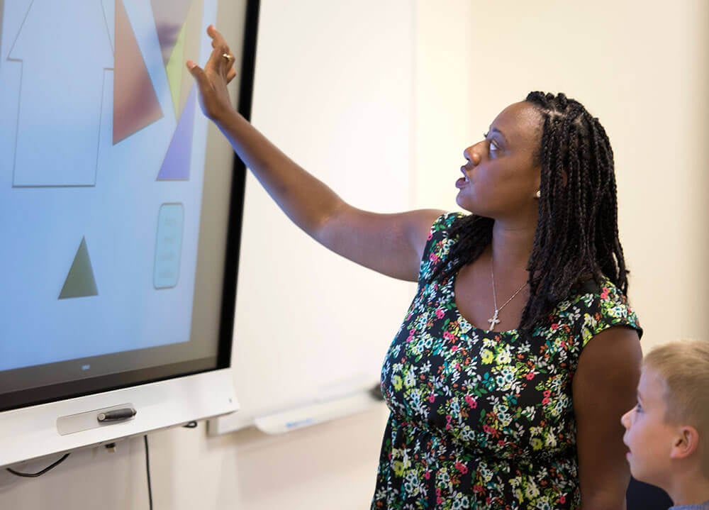 A School of Education graduate student gestures toward a large digital screen as an elementary student looks on