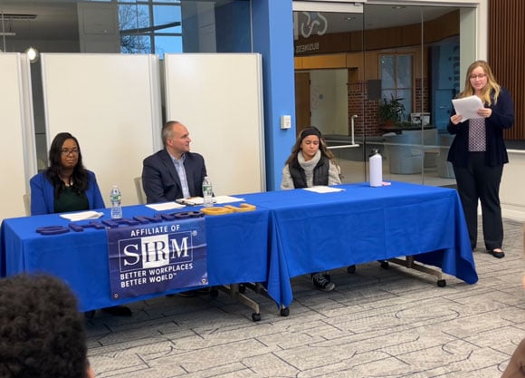 Panelists sit at a royal blue table as a Quinnipiac HR Student presents