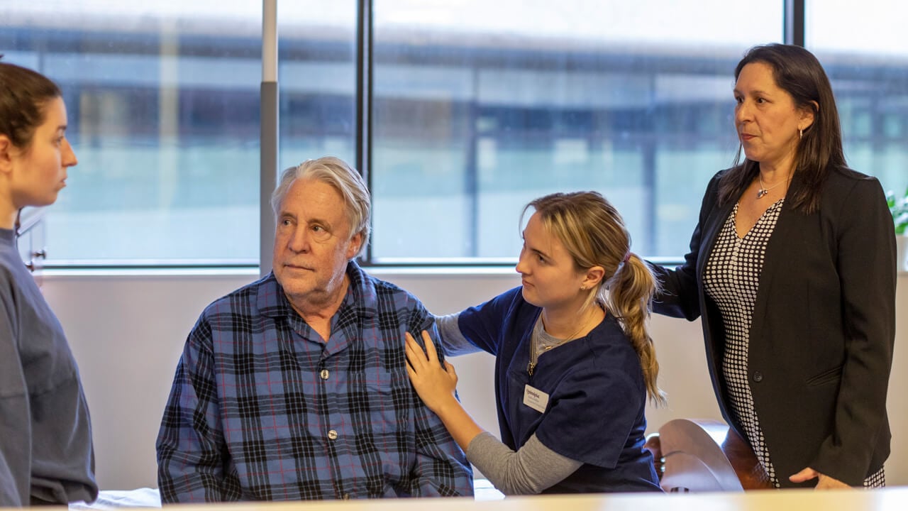 2 students work with a standardized patient with a professor watching