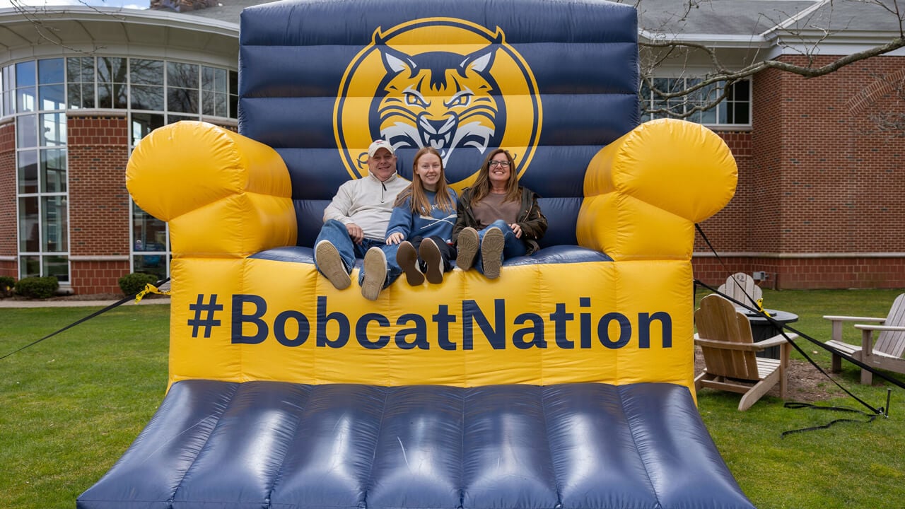 A new student sits with her family on a giant inflatable chair that says #BobcatNation on the quad