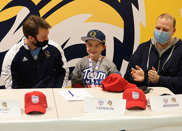 A boy smiling at the camera wearing a Team IMPACT shirt, seated next to two men. There are three red hats on the table in front of them.