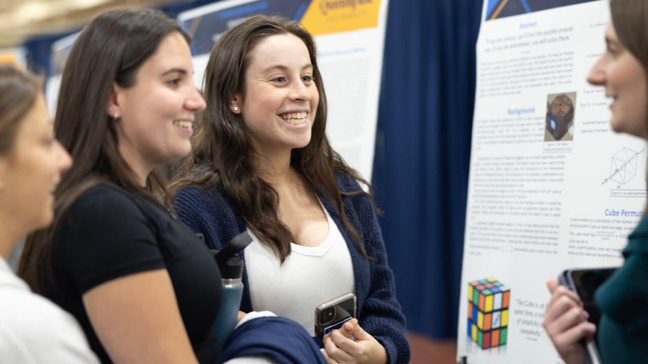 Quinnipiac student presents her research poster to two other students who are pictured smiling