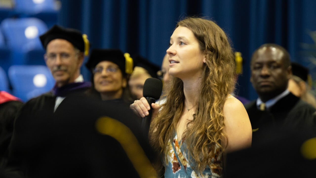 Cameron Chaplen holds a microphone and sings in front of the graduates