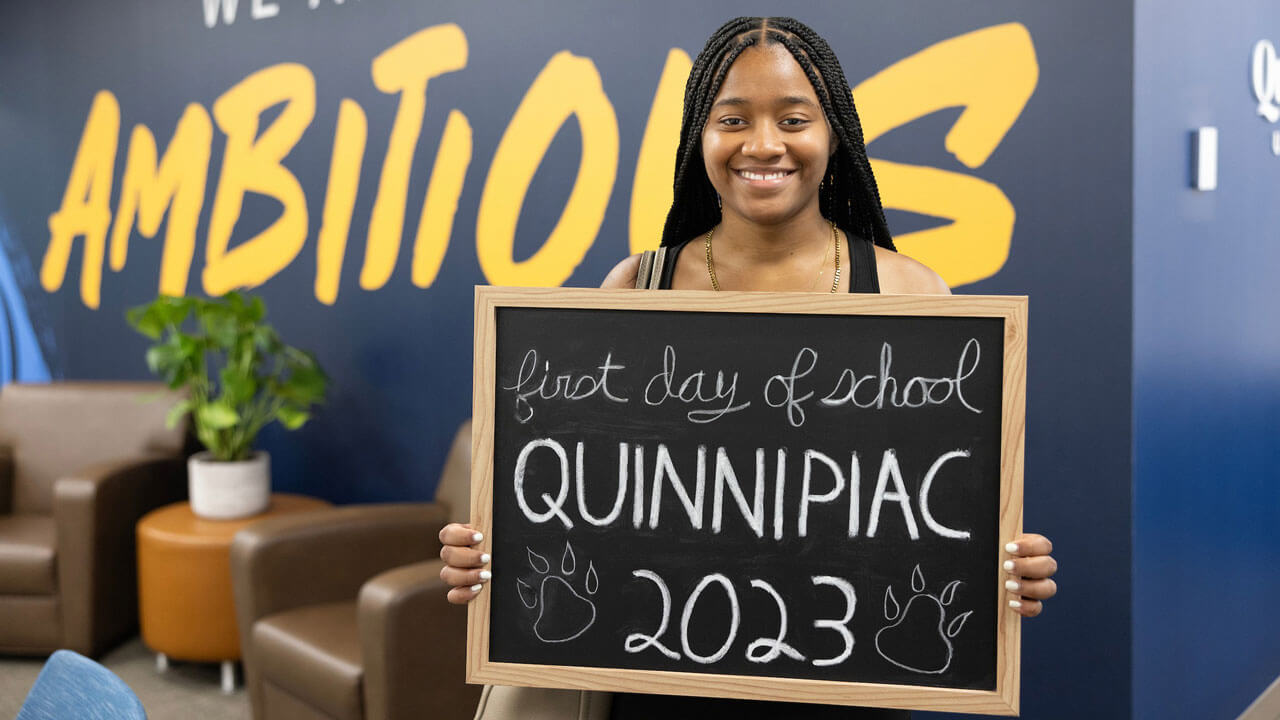 Student smiling with chalkboard in hand in front of bright yellow ambitious sign