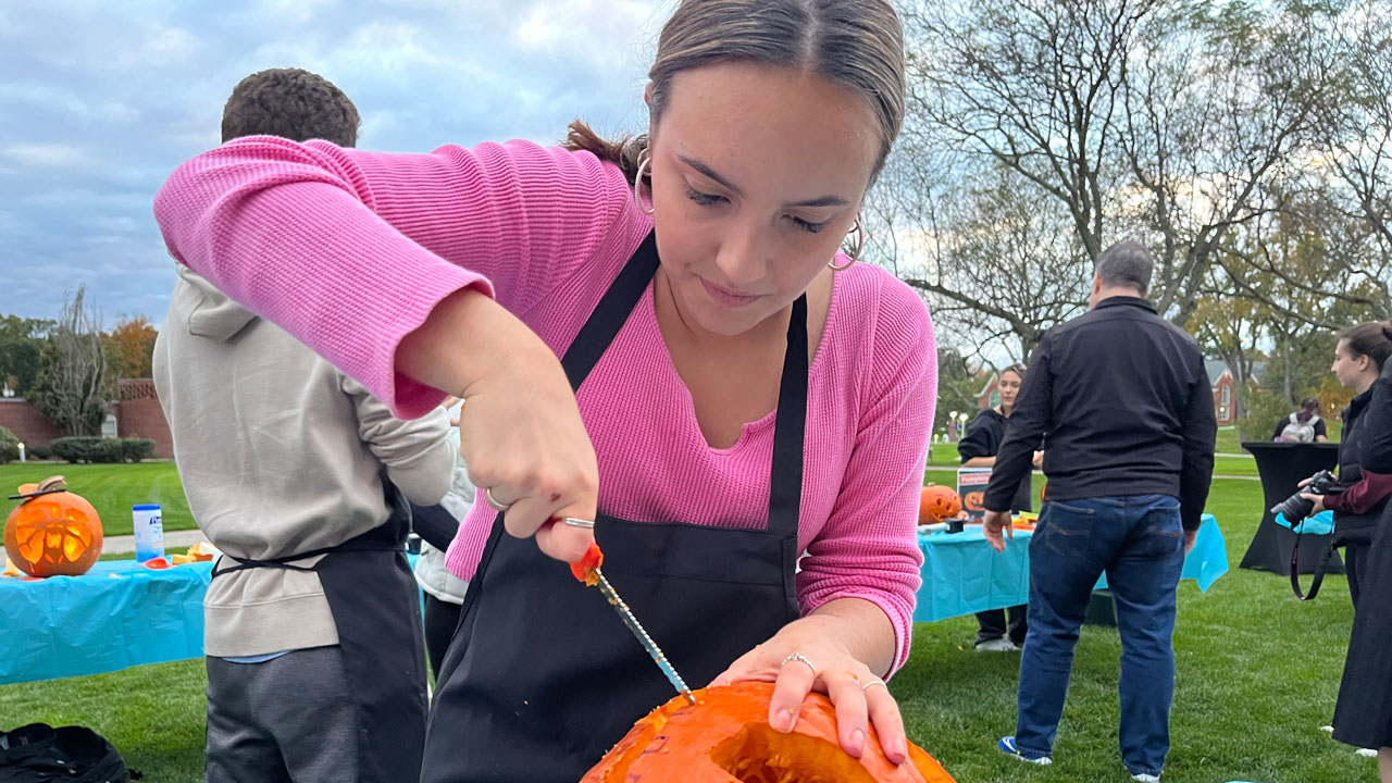 A female student wearing an apron and pink shirt concentrates on carving her pumpkin