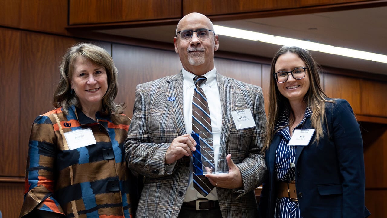 An awardee smiles holding a trophy next to two other professionals at the inaugural alumni association awards.