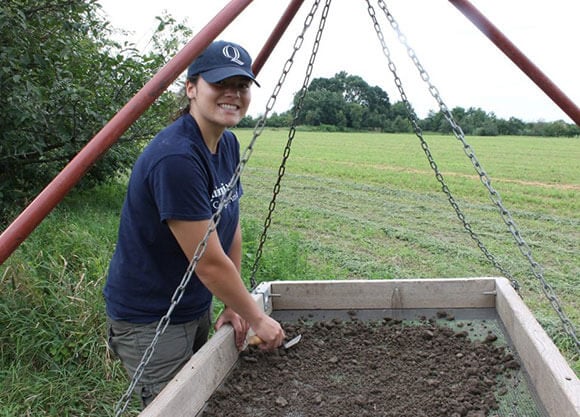 Erika Danella stands in a field working with a screen and trowel searching through dirt.