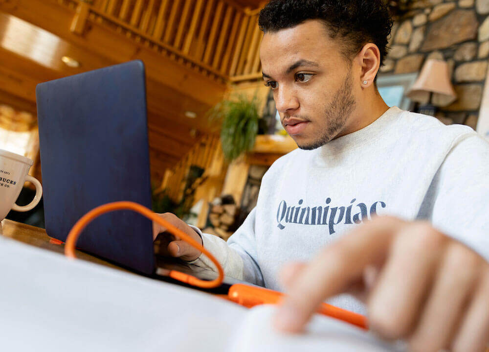 A student wearing a gray Quinnipiac sweater works on his laptop from home