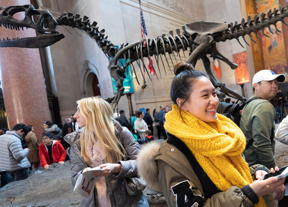 Quinnipiac students smile and stand in the Natural History museum in New York City with a T-rex skeleton in the background