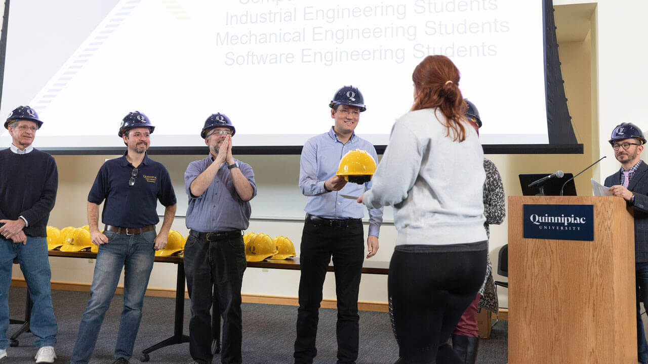 A faculty member passes a hard hat to a student