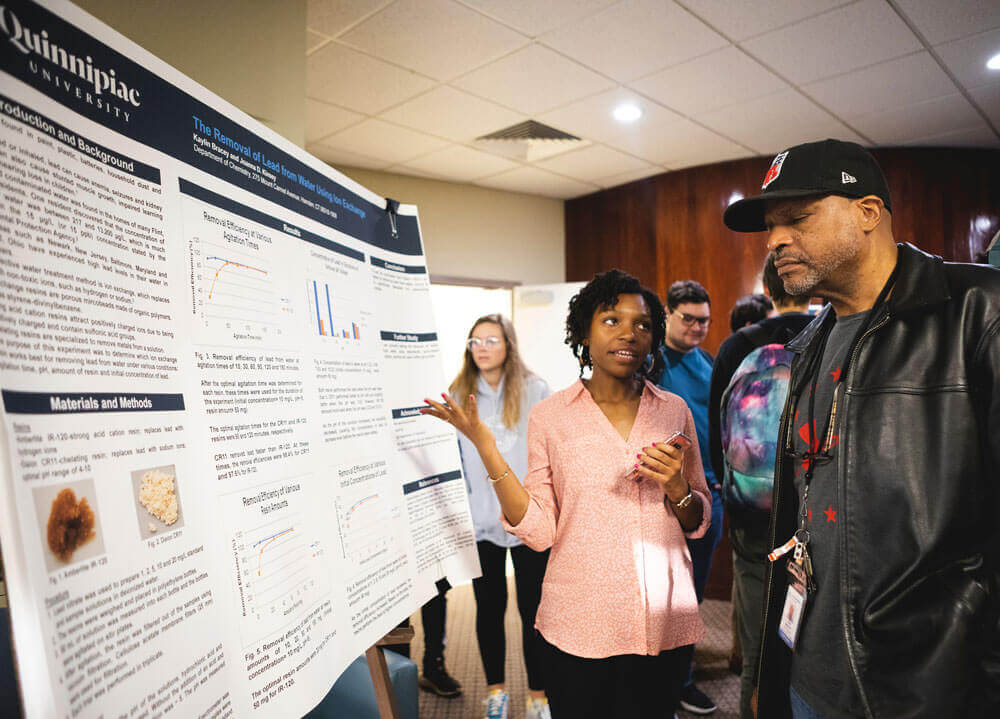 Kaylin Bracey, a Quinnipiac chemistry student, presents her research on a poster to a man at the QUIP-RS symposium.